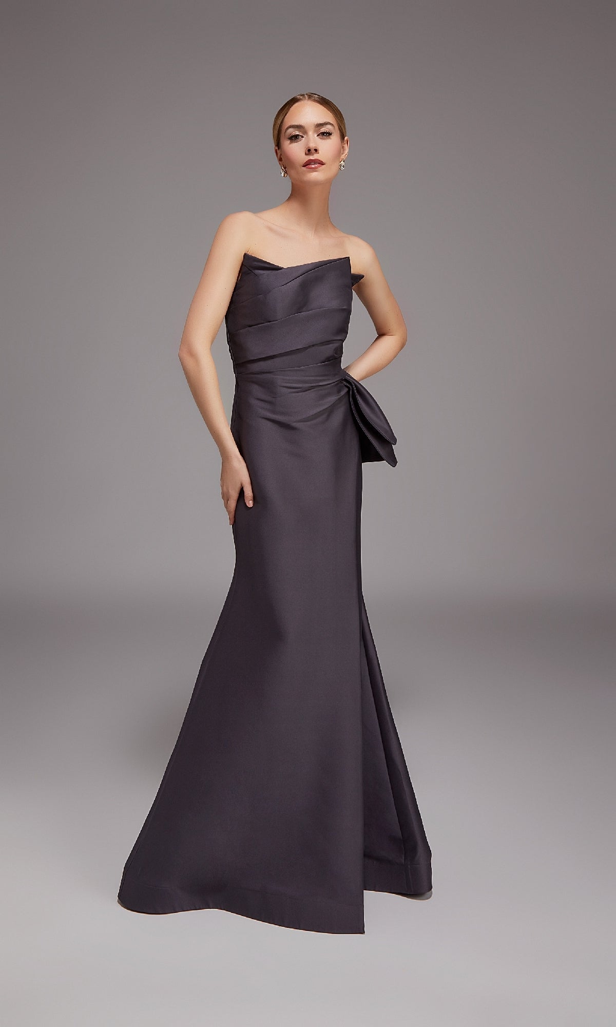 A woman poses in a studio wearing the Alyce 27806 Long Formal Dress, a strapless, floor-length charcoal gown with an elegant side bow detail.
